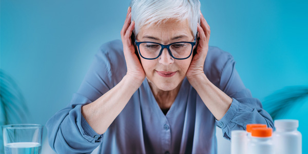 Eine Person sitzt an einem Tisch, stützt den Kopf mit beiden Händen und trägt ein langärmliges Oberteil; im Vordergrund stehen mehrere Medikamentenbehälter und ein Glas Wasser.