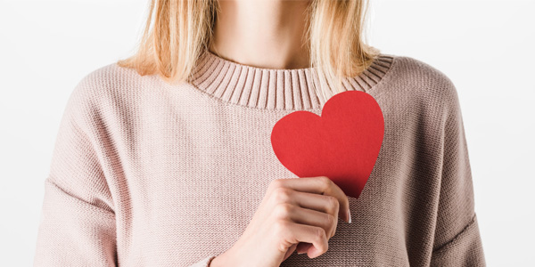 Oberkörper einer Person in einem hellen Strickpullover, die ein rotes, herzförmiges Papier vor der Brust hält.